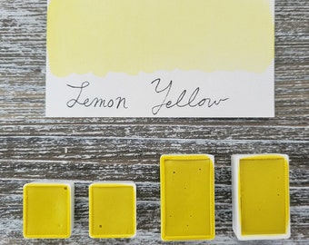 May include: A rectangular white card with a lemon yellow paint swatch and the handwritten text Lemon Yellow, accompanied by four yellow watercolor pans in two different sizes, arranged in a row below the card on a weathered wood surface.