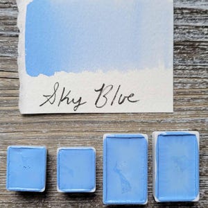 May include: A watercolor paint swatch labeled "Sky Blue" with a gradient of light blue. Below are three square and rectangular pans filled with sky blue watercolor paint. The pans are in white ceramic containers.