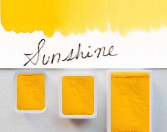 May include: Three rectangular white plastic pans filled with vibrant yellow watercolor paint. Above the pans, a swatch of yellow paint is applied to white paper with the word Sunshine written in black cursive script below the paint swatch.