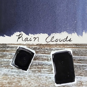 Pode incluir: Duas pastilhas retangulares de aquarela preenchidas com tinta azul escura, com a etiqueta "Rain Clouds" em itálico. Uma amostra de tinta azul escura está acima das pastilhas. O fundo é uma superfície de madeira clara e desgastada.