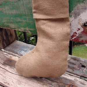 May include: A brown burlap stocking with a folded cuff and a loop at the top. The stocking is sitting on a wooden surface.