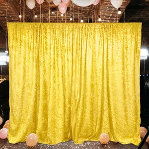 May include: A large, golden yellow velvet curtain backdrop. The fabric is heavily textured and draped, creating deep folds and shadows. The backdrop is set up for an event, with balloons and lights visible in the background.