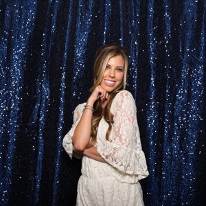 May include: A woman with long blonde hair is wearing a white lace dress. She is standing in front of a blue sequin backdrop.