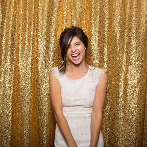 May include: A woman in a white lace dress smiles and laughs in front of a gold sequin backdrop.
