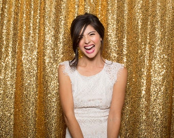 May include: A woman wearing a white lace dress smiles and laughs in front of a gold sequin backdrop.