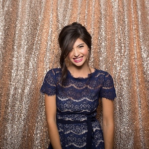 May include: A woman wearing a blue lace dress with a black belt, smiling and looking at the camera. She is standing in front of a gold sequin backdrop.