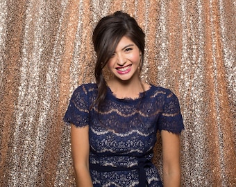 May include: A woman wearing a blue lace dress with a black belt, smiling and looking at the camera. She is standing in front of a gold sequin backdrop.