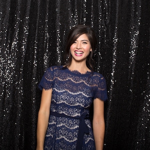May include: A woman wearing a blue lace dress with a black belt, smiling against a black sequin backdrop.