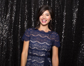May include: A woman wearing a blue lace dress with a black belt, smiling against a black sequin backdrop.