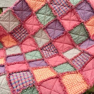 May include: A patchwork quilt in shades of pink, orange, green, purple, and beige. The quilt is made of squares and triangles, with a frayed edge. The quilt is displayed on a wooden bench. The quilt is made of various fabrics.