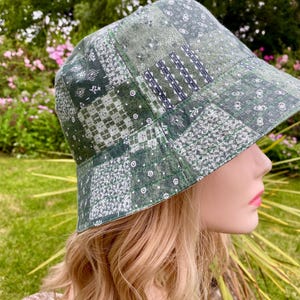 May include: A bucket hat featuring a patchwork design in various shades of green, white, and dark grey. The fabric displays multiple geometric and floral patterns stitched together, creating a textured appearance across the entire surface of the hat.