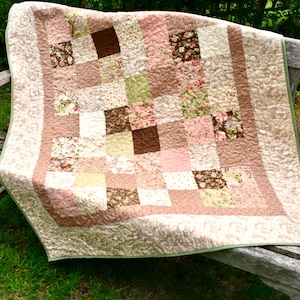 May include: A patchwork quilt featuring a variety of square fabric blocks in shades of pink, brown, cream, and sage green. The design includes solid colours and floral patterns, finished with a wide border and intricate overall quilting stitches.
