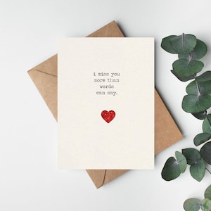 May include: A cream-colored greeting card with the text "i miss you more than words can say" and a red glitter heart. The card is on a brown envelope, with green leaves in the background.