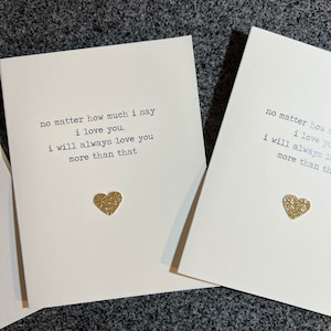 Może przedstawiać: Kremowe kartki okolicznościowe z napisem "no matter how much I say I love you, I will always love you more than that." Każda karta ma złote serce z brokatem. Kartki leżą na ciemnoszarej powierzchni.