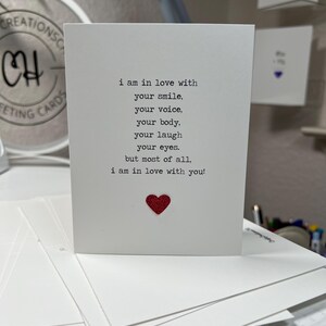 May include: A white greeting card with the text "i am in love with your smile, your voice, your body, your laugh, your eyes, but most of all, i am in love with you!" and a red glitter heart.