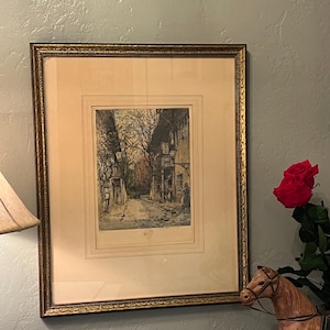 May include: Framed vintage print depicting a European street scene with buildings and figures. The print is in a gold-toned frame and is mounted on a cream-colored mat. A red rose and a decorative horse figurine are next to the framed print.