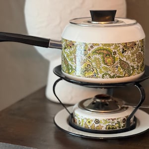 May include: A vintage fondue set with a white pot featuring a green and gold paisley pattern, black handle, and lid. The set includes a stand and a smaller burner, all on a dark wooden surface.