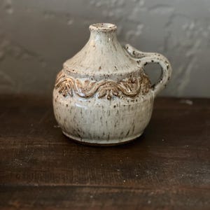 May include: A speckled, off-white ceramic pitcher with a handle and a decorative band of raised floral designs. The small jug has a rounded body and a narrow neck. The handle is small and curved. The pitcher is sitting on a dark wooden surface.