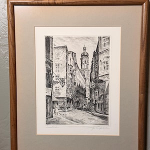 May include: A framed black and white etching depicting a narrow European street scene with tall buildings, a prominent church tower in the distance, and ornate hanging signs. The artwork is matted in a light tan border and enclosed in a wooden frame.