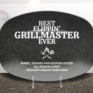 May include: A black oval-shaped platter with white text that reads "BEST FLIPPIN' GRILLMASTER EVER." Below the text is a graphic of a grill and utensils. The platter also includes the text "BOBBY, THANKS FOR KEEPING US FED ALL SEASON LONG! TAILGATE DREAM TEAM 2026."
