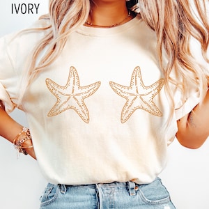 May include: Ivory-colored t-shirt featuring two starfish designs in a light brown color. The word "IVORY" is printed at the top. The shirt is a casual, short-sleeved top.