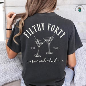 May include: Dark gray t-shirt with the text "FILTHY FORTY" above two martini glasses and the words "EST 1985" and "social club". The shirt has a vintage, worn look.