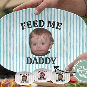 May include: An oval-shaped serving platter with a light blue and white striped background. The platter features the text "FEED ME DADDY" with a baby's face in the center. The platter is held by a person.