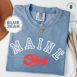 May include: A blue jean t-shirt with the word "MAINE" in white block letters and a red lobster illustration. The shirt is made of a soft, comfortable material.