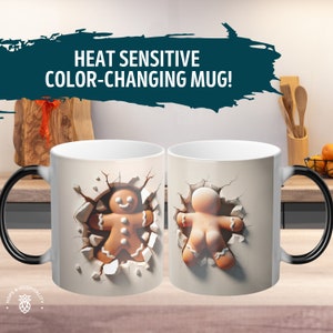 May include: A white ceramic mug with a black handle. The mug is a color-changing mug, and when hot liquid is poured into it, the image changes to reveal a gingerbread man breaking out of a wall. The mug has the text "HEAT SENSITIVE COLOR-CHANGING MUG!" printed on it.