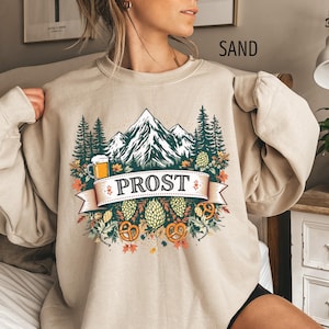 May include: Sand-colored sweatshirt with a mountain and forest graphic. The design includes a beer stein, pretzels, and the word "PROST" on a banner. The sweatshirt is a crew neck style.