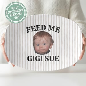 K&ouml;nnte beinhalten: Ovale Keramikplatte mit vertikalem Streifenmuster in Beige und Wei&szlig;. Die Platte zeigt das Gesicht eines Babys und die Worte "FEED ME" dar&uuml;ber und "GIGI SUE" darunter. Ein vollst&auml;ndig personalisiertes Andenken.