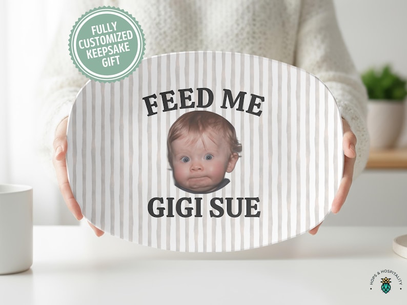 K&ouml;nnte beinhalten: Ovale Keramikplatte mit vertikalem Streifenmuster in Beige und Wei&szlig;. Die Platte zeigt das Gesicht eines Babys und die Worte "FEED ME" dar&uuml;ber und "GIGI SUE" darunter. Ein vollst&auml;ndig personalisiertes Andenken.