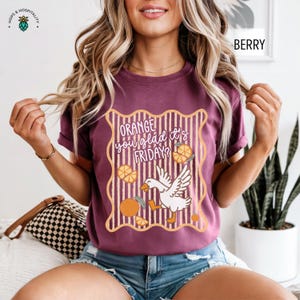 May include: A berry-colored t-shirt featuring a graphic with a white goose, orange slices, and vertical stripes inside a wavy border. The text reads, "ORANGE you glad it's FRIDAY?" The design uses orange, white, and dark brown colors on the fabric.