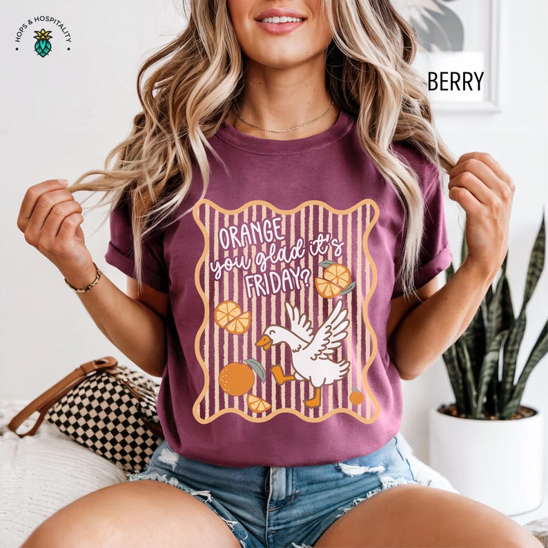 May include: A berry-colored t-shirt featuring a graphic with a white goose, orange slices, and vertical stripes inside a wavy border. The text reads, "ORANGE you glad it's FRIDAY?" The design uses orange, white, and dark brown colors on the fabric.