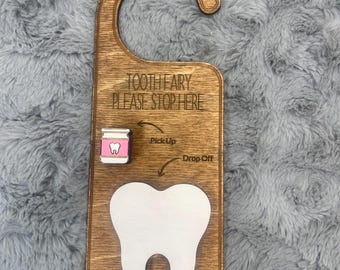 May include: Wooden door hanger with a tooth fairy theme. The brown hanger has a cutout for the doorknob and features the text "TOOTH FAIRY PLEASE STOP HERE". It includes a white tooth graphic and a pink tooth-shaped container.