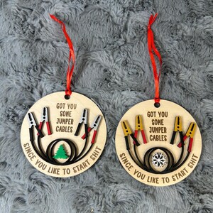 May include: Two wooden Christmas ornaments with red ribbons. Each ornament is round and features the text "GOT YOU SOME JUMPER CABLES SINCE YOU LIKE TO START SHIT." One has a green tree, the other a snowflake.