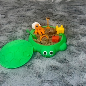 May include: A green turtle-shaped sandbox toy with a lid. Inside, there's sand and small toys, including a figure, shovel, bucket, and other accessories. The lid is open, revealing the play area. The toy is on a gray, fluffy surface.