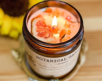 May include: A brown glass candle jar with a light orange wax candle inside. The candle has a dried citrus slice and other botanicals embedded in the wax. The jar has a label that reads "Botanical Garden - Eureka".