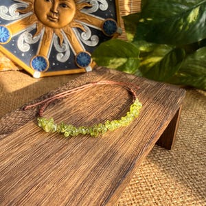 May include: A delicate bracelet featuring small, light green peridot gemstones strung on a thin, brown cord. The bracelet is displayed on a wooden surface, with a decorative sun-faced tile in the background. The gemstones have a natural, irregular shape.