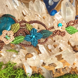 Peut inclure: Un bandeau macramé fait à la main avec une grosse pierre précieuse ovale bleue. Le bandeau est orné de macramé marron, de feuilles vertes et de petites fleurs beiges avec des centres turquoise. Le design est posé sur un fond de cristaux de quartz clairs.