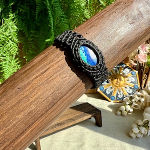Peut inclure: Bracelet en macramé noir orné d'une pierre ovale bleue et verte. Le bracelet est présenté sur un support en bois. Le fond comprend de la verdure et une tuile décorative en forme de soleil.