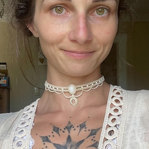 May include: A cream-colored lace choker necklace with a central oval-shaped pendant and a smaller, round pendant below. The necklace is worn on a person's neck. The person is wearing a white top with lace detailing.