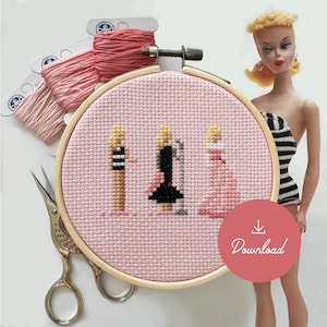 May include: A pink embroidery hoop with a cross-stitch design of three women in different outfits. The women are wearing a black and white striped dress, a black dress, and a pink dress. The hoop is surrounded by embroidery floss in various shades of pink.