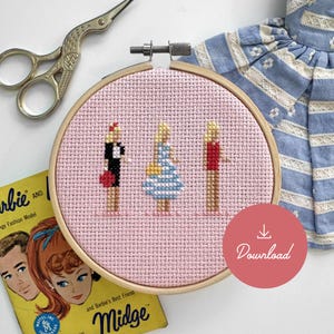 May include: A pink cross-stitch embroidery hoop with three figures of Barbie dolls. The dolls are wearing different outfits, including a red dress, a blue and white striped dress, and a red shirt. The hoop is on a yellow book with the text "Barbie and Barbie's Best Friend Midge".