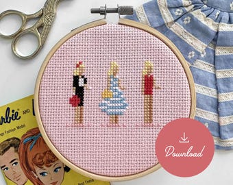 May include: A pink cross-stitch embroidery hoop with three figures of Barbie dolls. The dolls are wearing different outfits, including a red dress, a blue and white striped dress, and a red shirt. The hoop is on a yellow book with the text "Barbie and Barbie's Best Friend Midge".
