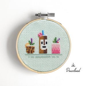 May include: A cross-stitch embroidery hoop with a light green background featuring a brown tiki, a pink cocktail, and a small potted plant. The embroidery is framed in a natural wood hoop.