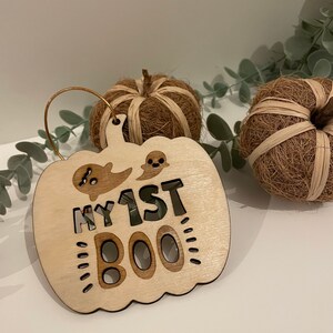 May include: A wooden pumpkin-shaped ornament with the text "My 1st Boo" and a ghost cutout. The ornament is light brown and has a gold string for hanging.