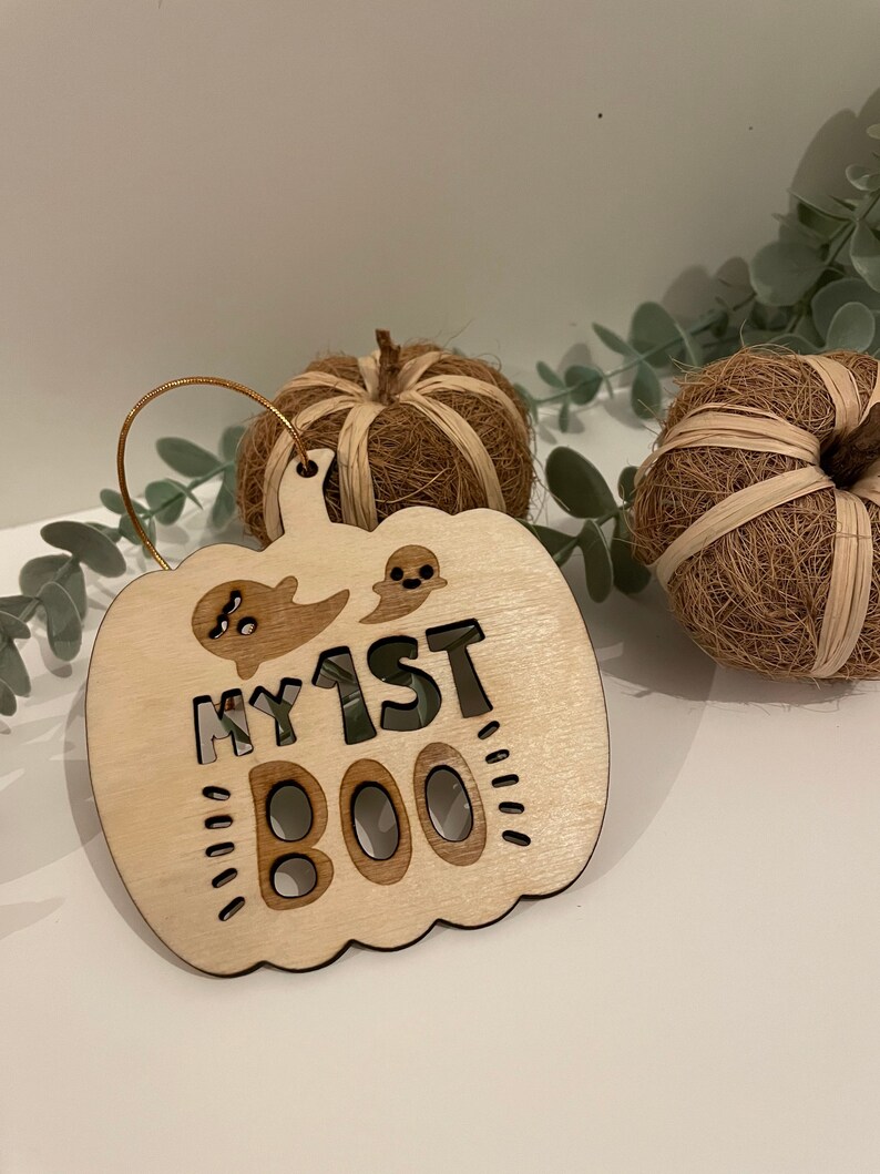 May include: A wooden pumpkin-shaped ornament with the text "My 1st Boo" and a ghost cutout. The ornament is light brown and has a gold string for hanging.