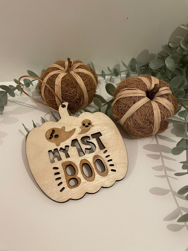 May include: Wooden pumpkin-shaped ornament with the text "My 1st Boo" and a ghost cutout. The ornament is light brown and has a string for hanging.
