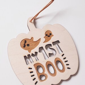 May include: A wooden pumpkin-shaped ornament with the text "MY 1ST BOO" and two ghost cutouts. The ornament is painted in a light wood color and has a brown string for hanging.